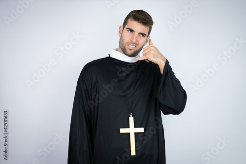 Young hispanic man wearing priest uniform standing over white background confused doing phone gesture with hand and fingers like talking on the telephone