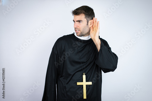 Young hispanic man wearing priest uniform standing over white background surprised with hand over ear listening an hearing to rumor or gossip