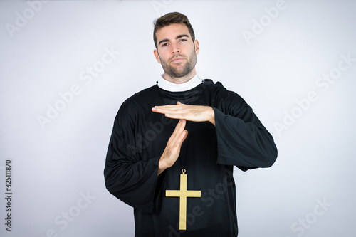 Young hispanic man wearing priest uniform standing over white background Doing time out gesture with hands, frustrated and serious face