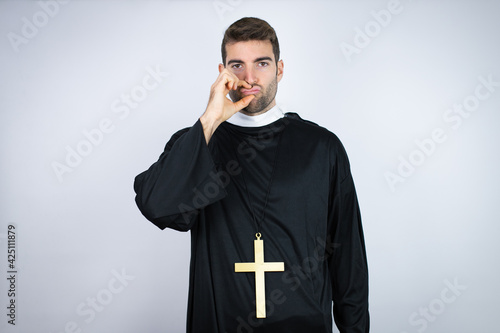 Young hispanic man wearing priest uniform standing over white background mouth and lips shut as zip with fingers. Secret and silent, taboo talking