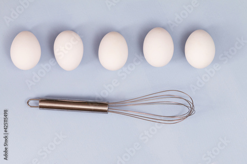 group of white chicken eggs on light blue background with stainless steel whisk   empty space for text 