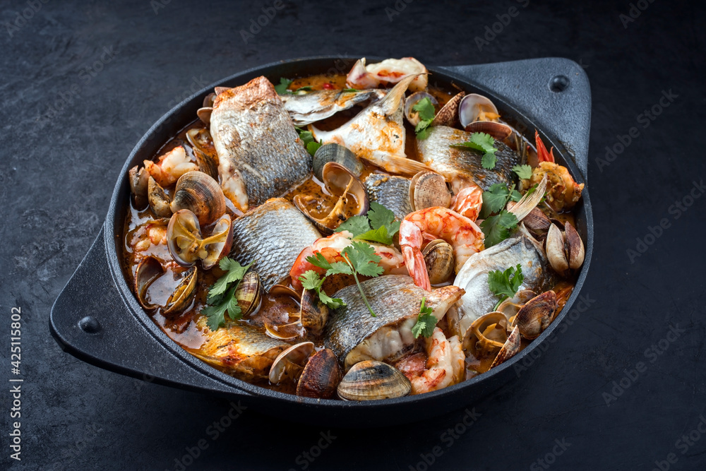 Modern style traditional Spanish seafood zarzuela de pescado with fish, king prawns and venus clams served in clear sauce as close-up in design pot