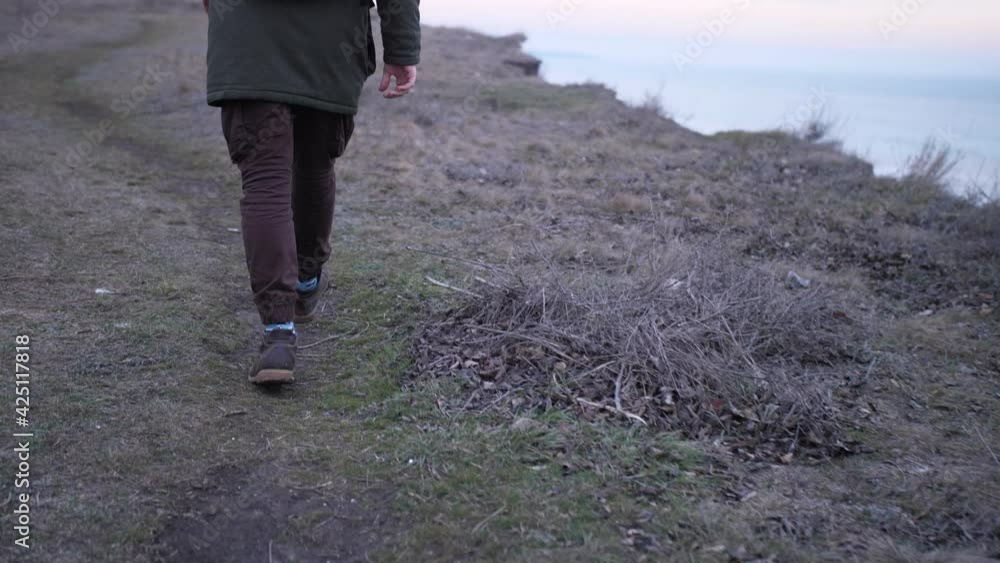 Back view a legs of man walking a dirty green hills above the sea. Lonely man solo trekking rugged terrain with depressive view at late autumn. Back view a legs of man walking a dirty green hills above the sea. Lonely man solo trekking rugged terrain with depressive view at late autumn.