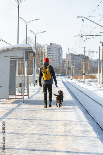 a Caucasian European man in a dark jacket and a medical mask with a Labrador retriever dog waits for a train at the railway station in winter, on a snowy day