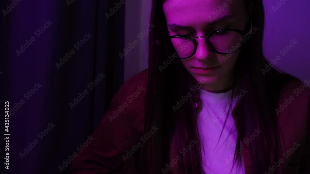 Focused young caucasian girl working on laptop sit on bed at home. Freelance girl in eyeglasses with reflections using computer in neon red light. Freelance business woman with notebook at night Focused young caucasian girl working on laptop sit on bed at home. Freelance girl in eyeglasses with reflections using computer in neon red light. Freelance business woman with notebook at night
