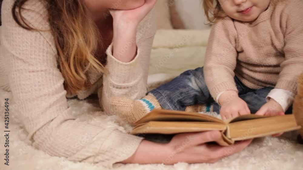 A young mother reads a book with her daughter. A woman and a girl play, laugh, leaf through the pages. Love caring home comfort family evening The child learns to read and talk. Beige bed interior Lie A young mother reads a book with her daughter. A woman and a girl play, laugh, leaf through the pages. Love caring home comfort family evening The child learns to read and talk. Beige bed interior Lie