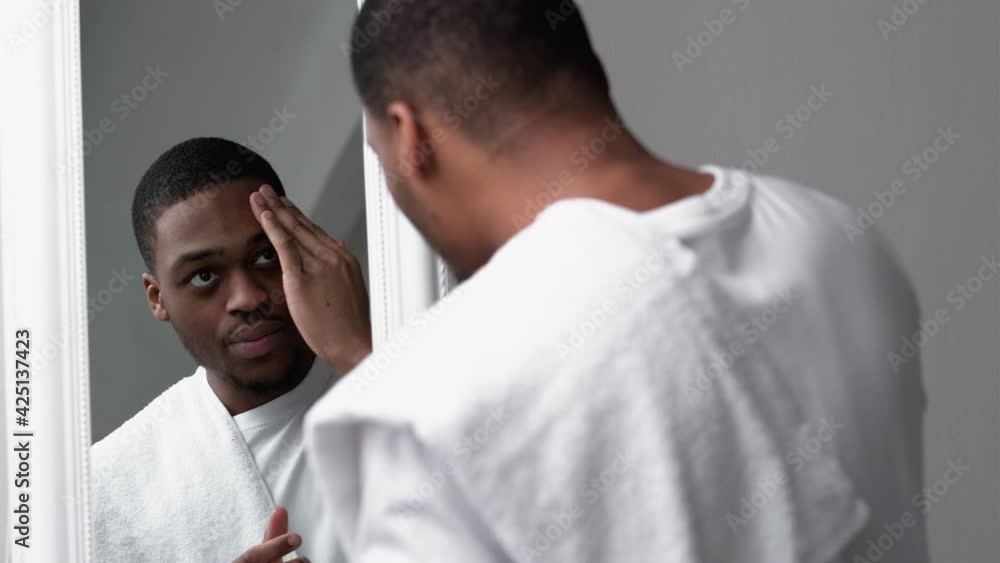 Man skincare. Acne treatment. Morning routine. Confident African guy in white t-shirt with towel touching face after cleansing looking in mirror. Man skincare. Acne treatment. Morning routine. Confident African guy in white t-shirt with towel touching face after cleansing looking in mirror.