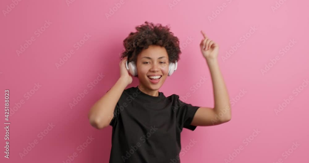 Positive dark skinned curly woman enjoys favorite audio track keeps hands on wireless headphones smiles broadly dressed in casual black t shirt isolated over pink background dances to pop music Positive dark skinned curly woman enjoys favorite audio track keeps hands on wireless headphones smiles broadly dressed in casual black t shirt isolated over pink background dances to pop music