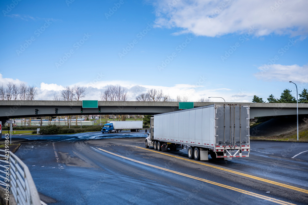 Big rigs semi trucks with semi trailers transporting cargo driving towards each other on the wide road intersection with overpass bridge road