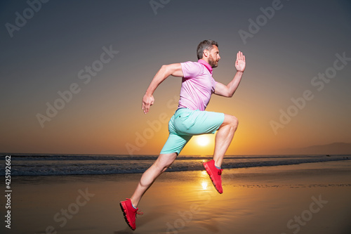 Athletic young man running in the nature. Fit man fitness model working outdoor by sea beach. Dynamic run movement.