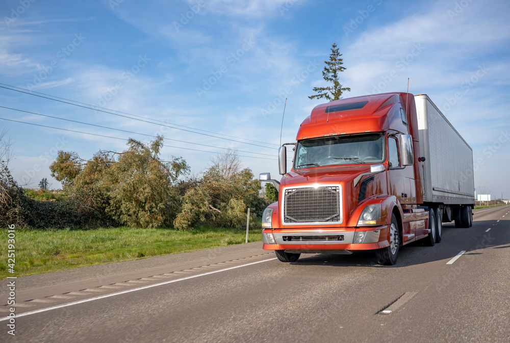 Bright brown big rig semi truck transporting cargo in refrigerator semi trailer driving on the multiline highway road with one way traffic direction