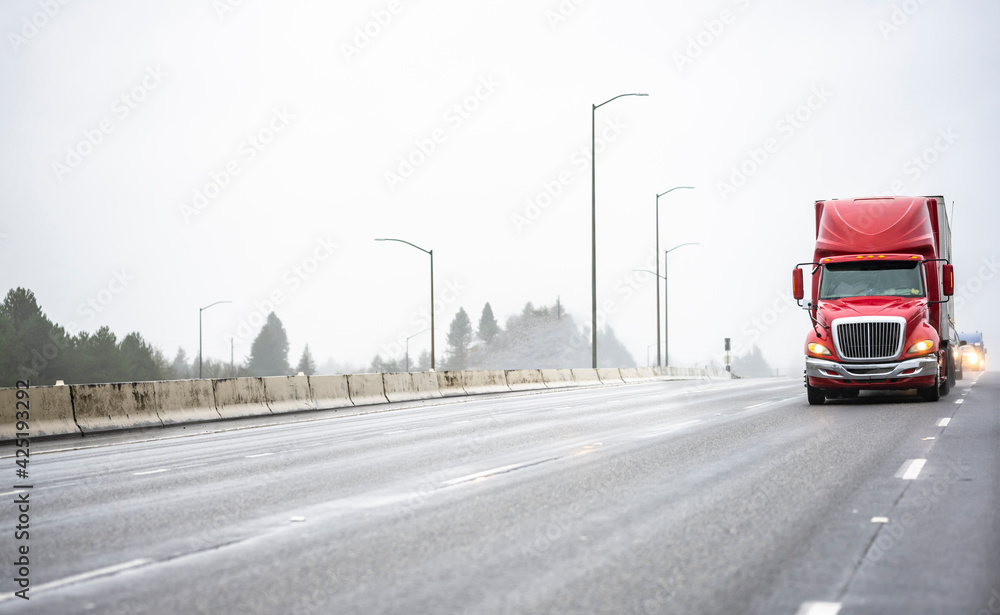 Bright red big rig bonnet semi truck transporting cargo in dry van semi trailer running on the wide multiline wet highway road with raining weather condition