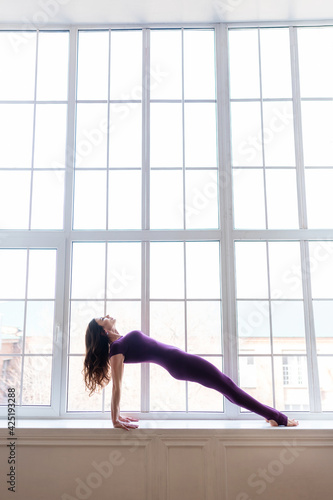 A beautiful dark-haired girl in a sports purple jumpsuit performs yoga asanas. Healthy lifestyle, sports.