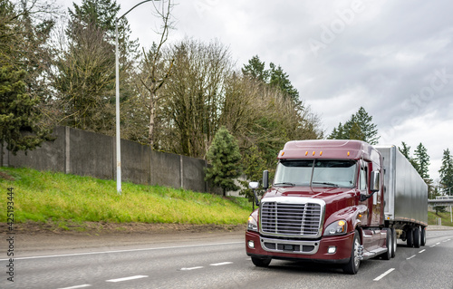 Burgundy low cab big rig semi truck transporting cargo in low profile covered semi trailer running on the multiline highway road with hill  and fence on the side