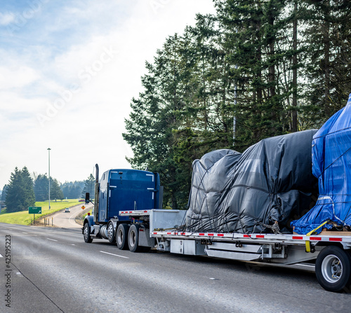 Huge blue classic big rig semi truck transporting covered heavy commercial cargo on step down semi trailer running on the wide highway road