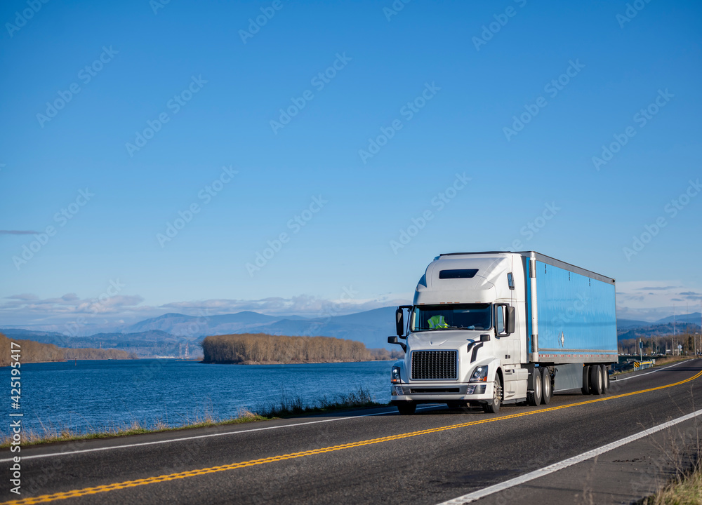 Industrial powerful white big rig semi truck transporting cargo in dry van semi trailer running on the narrow road along the Columbia River. Industrial powerful white big rig semi truck transporting cargo in dry van semi trailer running on the narrow road along the Columbia River.