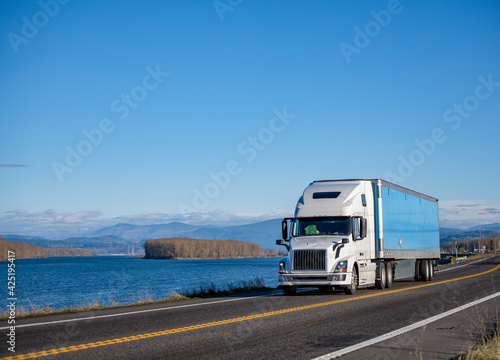 Industrial powerful white big rig semi truck transporting cargo in dry van semi trailer running on the narrow road along the Columbia River.