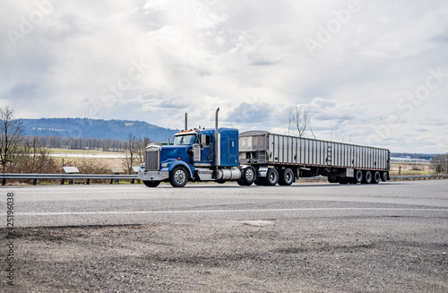 Powerful classic blue big rig American semi truck transporting cargo in low profile long bulk semi trailer running on the road in Columbia Gorge