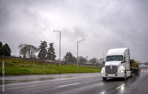 Powerful white big rig American bonnet semi truck transporting sling tightened cargo on flat bed semi trailer driving on the slippery wet road at storm rain condition weather
