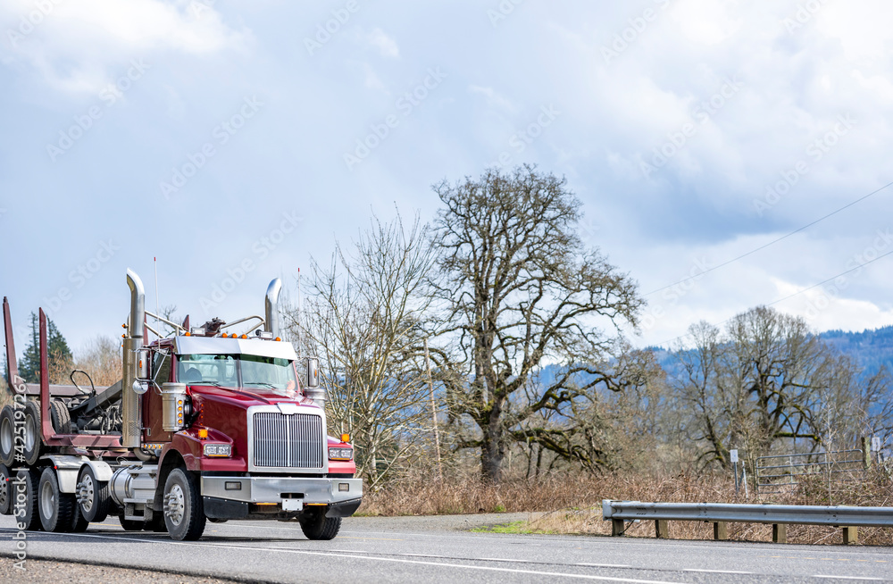 Red day cab big rig semi truck tractor transporting empty semi trailer for carry wood logs driving on the local road with trees on the side