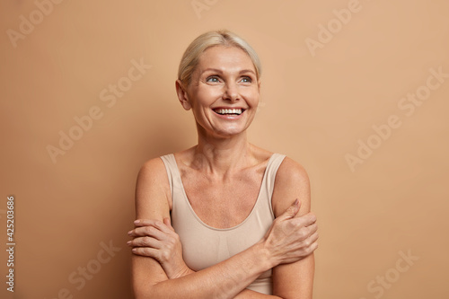 Attractive positive wrinkled fifty years old woman looks gladfully above keeps arms foded has well cared complexion healthy skin white teeth isolated over brown background. Beauty and age concept