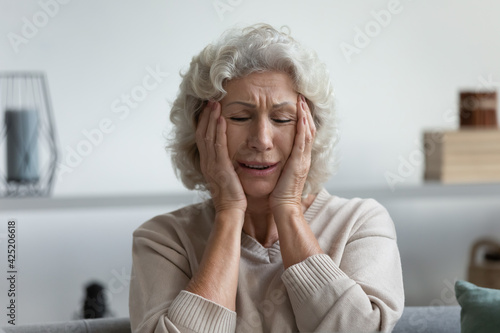 Upset exhausted mature 60s woman suffering from headache or painful migraine at home. Stressed frustrated elderly lady feeling dizziness, holding head, touching temples with pain grimace. Head shot