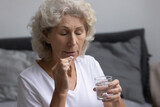 Just awaken senior lady holding morning daily dose of meds and glass of water, sitting on bed with closed eyes. Mature elder woman taking pills against memory loss, stress and mental disorder