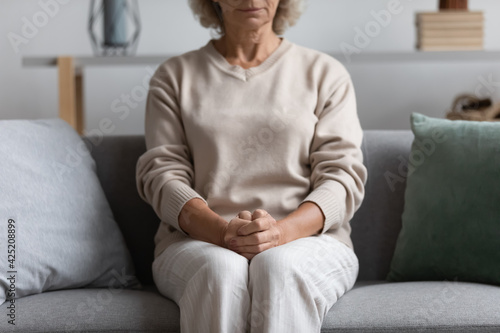 Worried concerned senior 60s lady keeping clasped hands on lap in anxious position, sitting on couch at home, expecting important news. Cropped shot of wrists, arms and knees of mature woman. Close up
