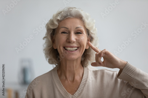 Portrait of happy senior woman pointing finger at her ear, looking at camera and smiling. Excited mature 60s lady enjoying hearing ability after deafness therapy. Deaf patient communication. Head shot photo