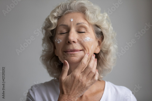 Relaxed elderly woman with closed eyes taking skincare treatment, applying anti age lotion, moisturizer cream on dry face skin with wrinkles. Senior beauty care, cosmetology concept. Close up portrait