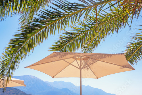 Beach vacation concept. Beach umbrella and  leaves of palm trees against blue sky and mountains on sunny day. Montenegro