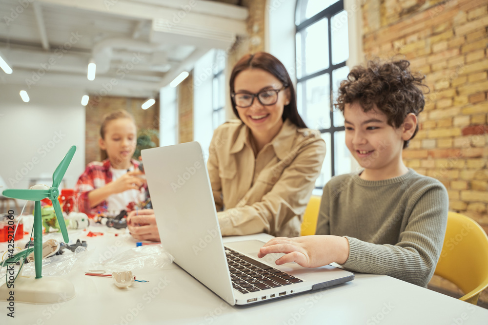 Young female teacher in glasses using laptop in classroom, showing scientific robotics video to little boy Young female teacher in glasses using laptop in classroom, showing scientific robotics video to little boy