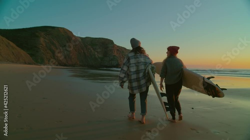 Two women walk with surfboards on empty epic beach towards surfing spot. Sunrise or sunset amazing views. Camera follows casual female surfers. Two friends have fun at beach