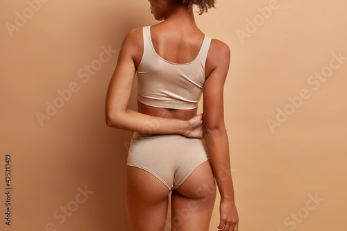 Back view of slim dark skinned woman with perfect buttocks wears cropped top and panties isolated over brown background. Health and beauty concept