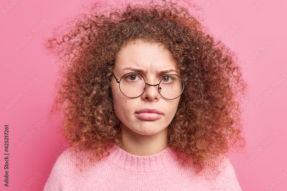 Close up portrait of dissatisfied curly haired woman looks angrily at camera smirks face wears transparent round spectacles cassual jumper isolated over pink background. Negative emotions concept Close up portrait of dissatisfied curly haired woman looks angrily at camera smirks face wears transparent round spectacles cassual jumper isolated over pink background. Negative emotions concept