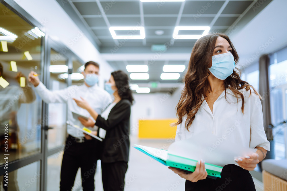 Young business woman in protective face mask an office building hallway. Teamwork of Business people during pandemic of coronavirus. Covid-19. Young business woman in protective face mask an office building hallway. Teamwork of Business people during pandemic of coronavirus. Covid-19.