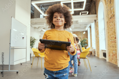 Portrait of cute little boy with afro hair smiling while holding and using tablet pc, standing in a classroom during STEM lesson photo