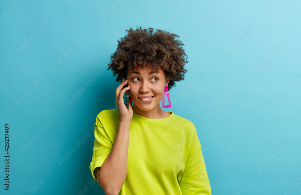 Beautiful curly haired woman talks casually with friend holds smartphone near ear bites lips gazes right enjoys good quality of device wears casual t shirt isolated on blue wall. Communication Beautiful curly haired woman talks casually with friend holds smartphone near ear bites lips gazes right enjoys good quality of device wears casual t shirt isolated on blue wall. Communication