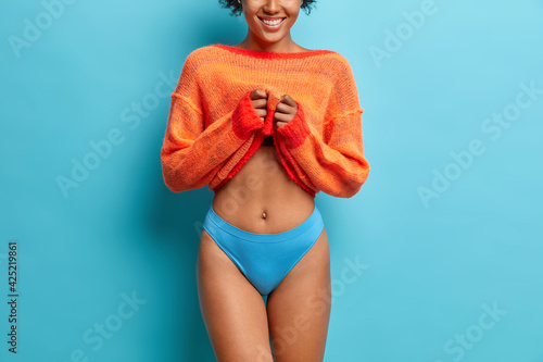 Faceless woman with toothy smile flat belly has beautiful body shape wears casual jumper shows her long slender legs isolated over blue background photo