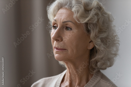 Pensive confused elderly 70s lady lost in thoughts, looking away at window. Close up portrait of senior pensioner woman forgetting things, suffering from memory loss, dementia, Alzheimer disease