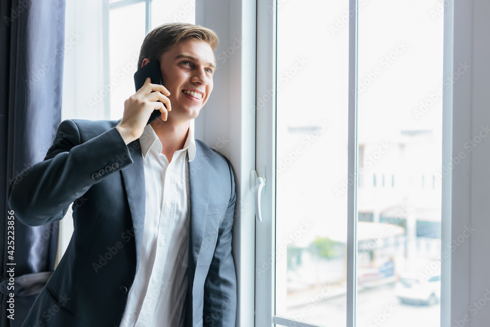 Businessman making phone call Standing near window in office at workplace Happy handsome business man in suit speaking on smartphone with client for sell goods Business Financial Communication concept Businessman making phone call Standing near window in office at workplace Happy handsome business man in suit speaking on smartphone with client for sell goods Business Financial Communication concept