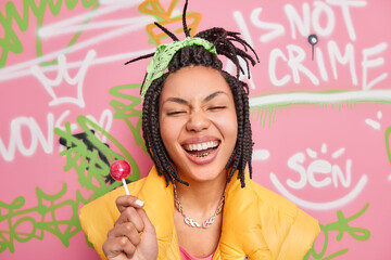 Trendy cheerful hipster girl smiles broadly holds lollipop has fun with teenagers of same age wears yellow vest poses against colorful graffiti wall. Gen z generation. Youth lifestyle concept