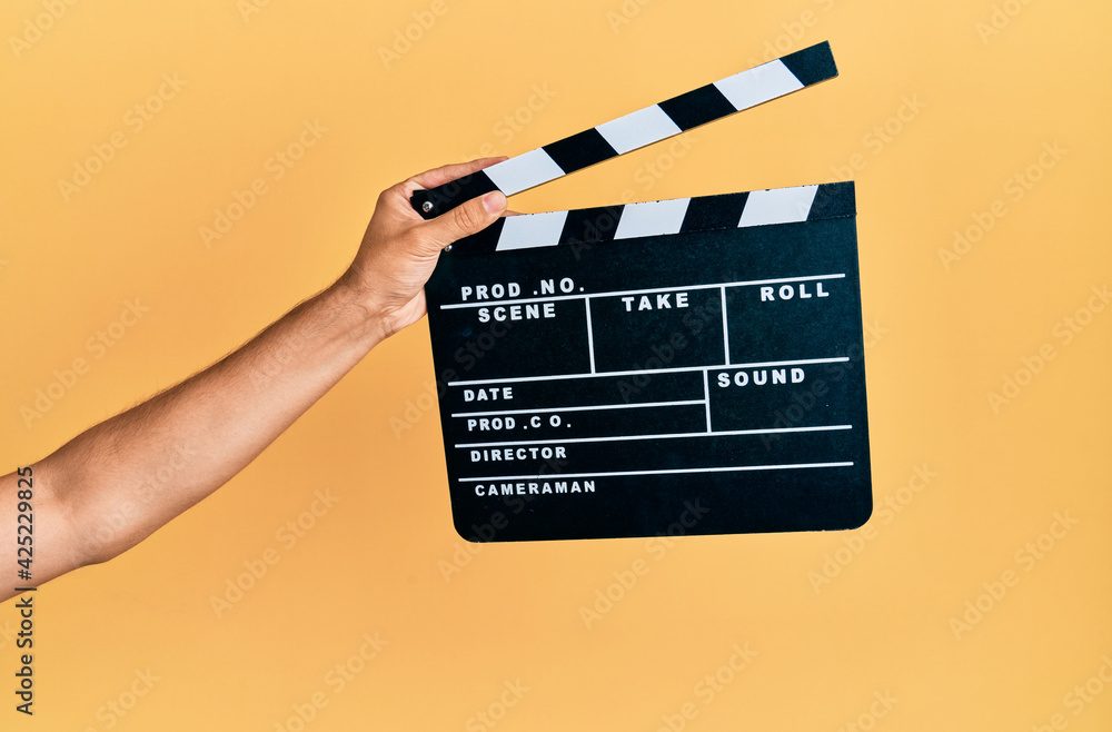 Hand of hispanic man holding movie clapboard over isolated yellow background.