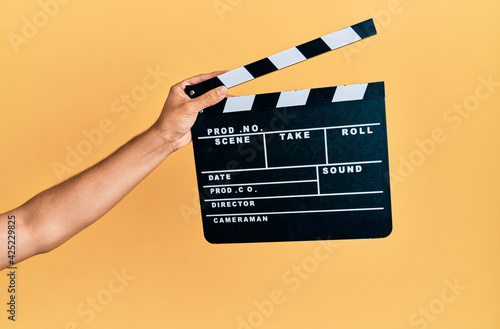 Hand of hispanic man holding movie clapboard over isolated yellow background. photo