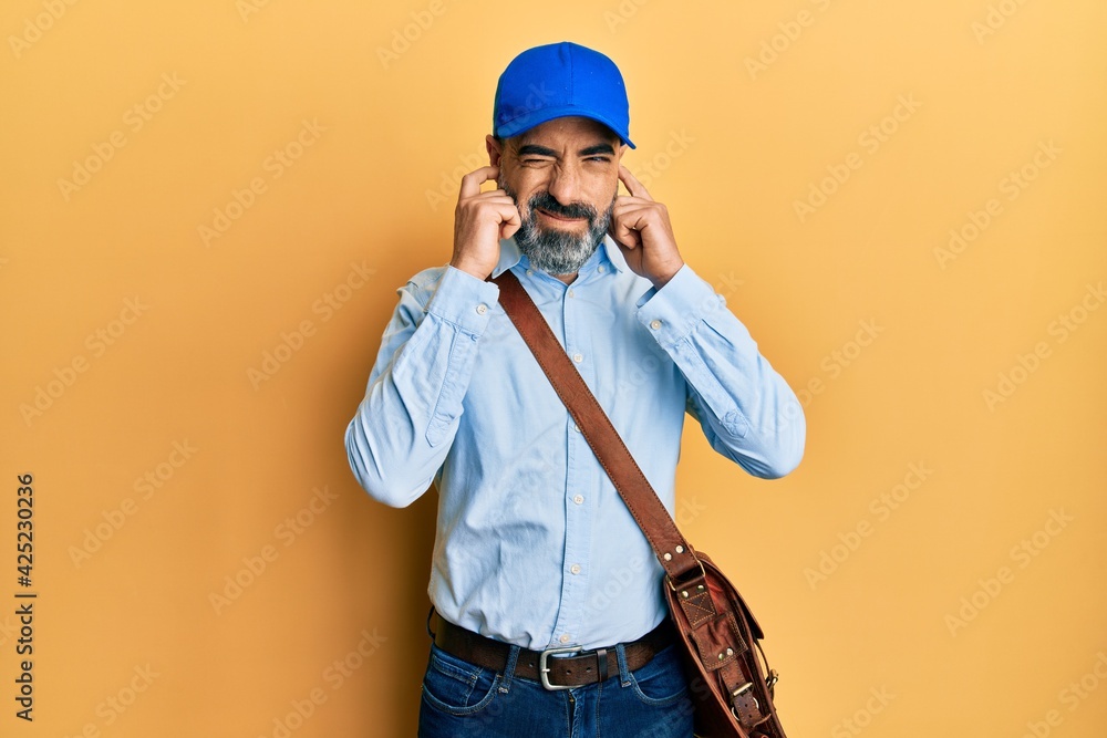 Middle age man with beard and grey hair wearing delivery courier cap covering ears with fingers with annoyed expression for the noise of loud music. deaf concept.