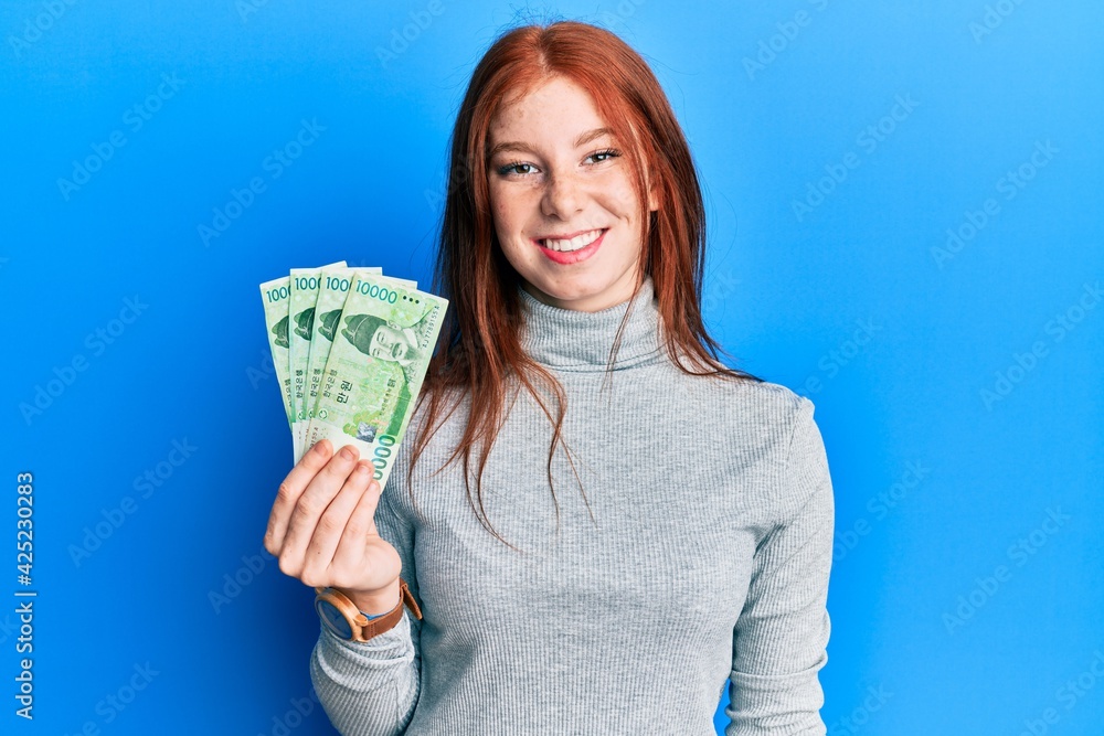 Young red head girl holding 10000 south korean won banknotes looking positive and happy standing and smiling with a confident smile showing teeth Young red head girl holding 10000 south korean won banknotes looking positive and happy standing and smiling with a confident smile showing teeth