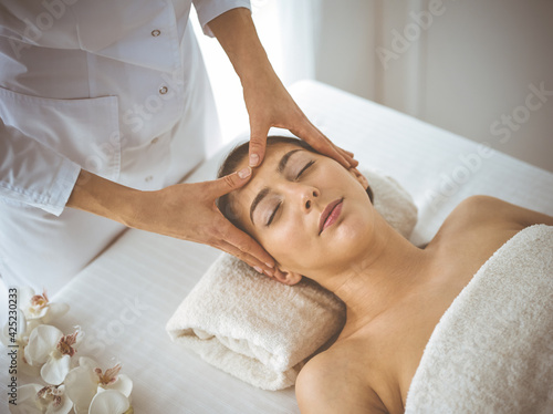 Beautiful brunette woman enjoying facial massage with closed eyes comfortable and blissful. Relaxing treatment in medicine and spa center concepts