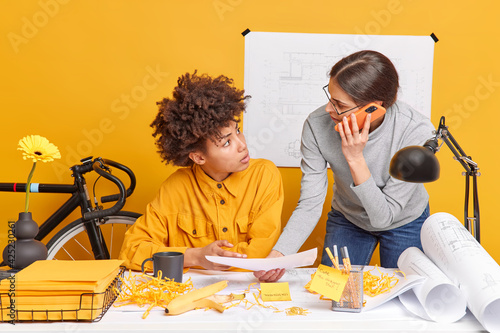 Multiracial women architects discuss future engineering project does paperwork pose at dekstop create blueprints isolated over yellow background have informal meeting at office. Collaboration