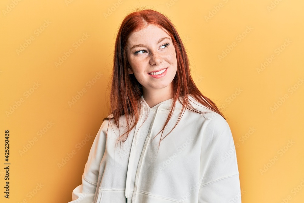 Young red head girl wearing casual sweatshirt looking away to side with smile on face, natural expression. laughing confident.
