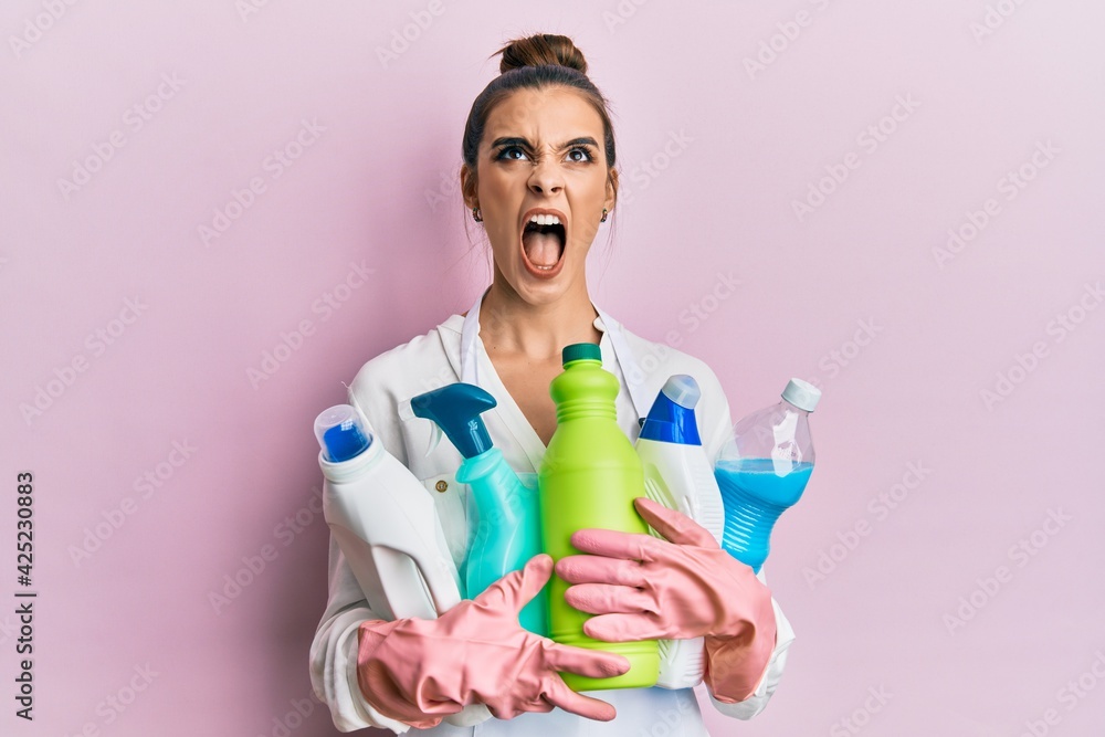 Beautiful brunette young woman wearing cleaner apron holding cleaning products angry and mad screaming frustrated and furious, shouting with anger looking up. Beautiful brunette young woman wearing cleaner apron holding cleaning products angry and mad screaming frustrated and furious, shouting with anger looking up.